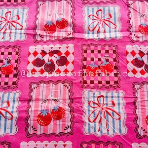 May include: Pink fabric featuring a repeating pattern of framed cherries, bows, and checkered designs. The cherries are red with green stems, and the bows are red and white. The fabric has a vintage, cottagecore aesthetic.