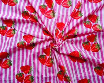 May include: Pink and white striped fabric with a pattern of red strawberries. The strawberries are whole and sliced, with green stems and leaves. The fabric is folded, creating a textured appearance.