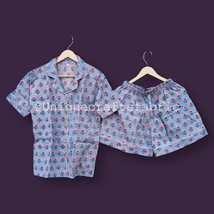 May include: A light blue pajama set with a floral pattern in pink and white. The set includes a short-sleeved button-down shirt and matching shorts. The edges of the shirt and shorts are trimmed with white piping. The text "@uniquecraftsfabric" is visible.