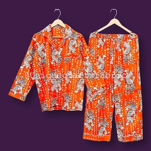 May include: Orange pajama set with a white tiger print. The set includes a button-down shirt and matching pants. The pajamas are hanging on wooden hangers against a purple background.