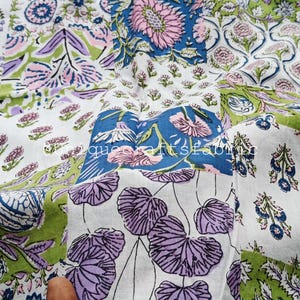 Op de afbeelding: Patchworkstof met diverse bloemen- en bladmotieven in blauw, groen, roze en paars. De stof heeft een witte achtergrond en de tekst "que craft fabric".