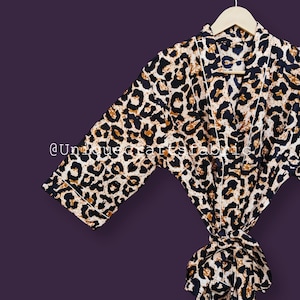 May include: A brown and black leopard print satin robe with a white trim and a tie belt. The robe is hanging on a wooden hanger against a purple background.