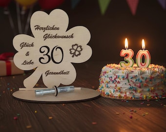 以下が含まれることがあります： 木製のクローバー型の誕生日飾りで、数字の30とドイツ語のテキストが書かれています。 30本のろうそくが立っている小さなケーキが飾りの隣にあります。 ケーキはカラフルなスプリンクルで飾られています。 背景には風船とギフトボックスがあります。