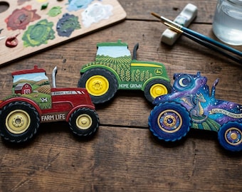 Könnte beinhalten: Drei dekorative Traktoraussparungen. Einer ist rot mit dem Text "FARM LIFE", ein anderer ist grün mit dem Text "HOME GROWN" und der dritte ist blau mit einem Drachen und dem Text "GALACTIC FARMER". Die Traktoren haben bemalte Details und befinden sich auf einer Holzoberfläche.
