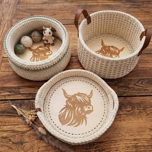 Könnte beinhalten: Drei handgefertigte Häkelkörbe mit Holzunterseiten, die ein Highland-Kuh-Design zeigen. Die Körbe sind in verschiedenen Größen erhältlich, mit cremefarbenem Garn und braunen Ledergriffen. Ein Korb enthält Wollknäuel und eine kleine Kuhfigur.