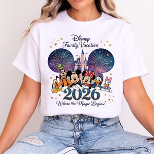 May include: A white t-shirt featuring a graphic of Disney characters including Mickey Mouse, Minnie Mouse, Goofy, Pluto, Donald Duck, and Daisy Duck in front of a castle and fireworks. The text reads Disney Family Vacation 2026 Where the Magic Begins.
