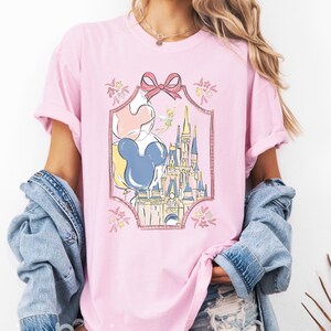 Peut inclure: Un t-shirt rose clair avec une impression graphique représentant un château, un groupe de ballons et une petite fée dans un cadre décoratif surmonté d'un nœud. Le texte LIGHT PINK est visible dans le coin supérieur droit de l'image.
