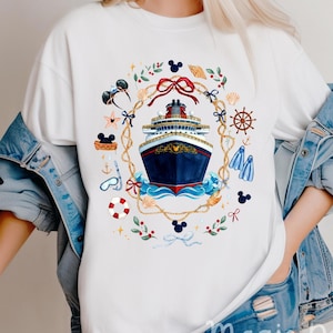 Peut inclure: Un t-shirt blanc avec un motif central représentant un navire de croisière bleu foncé entouré d'une couronne de corde. Le design inclut des éléments nautiques comme une barre de navire, une bouée, un tuba, des coquillages et des oreilles de Mickey.