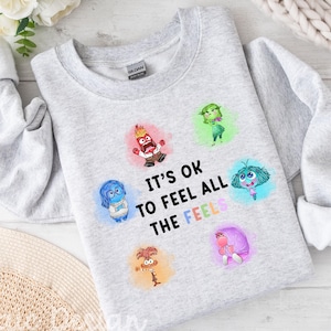 May include: Light grey sweatshirt featuring the phrase "It's ok to feel all the feels" and cartoon characters. The long-sleeved, crew-neck top displays characters in red, blue, green, and purple. The word "ASH" is printed in black.