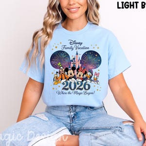 May include: A light blue t-shirt featuring a graphic of Disney characters including Mickey Mouse, Minnie Mouse, Donald Duck, Daisy Duck, Goofy, and Pluto in front of a castle with fireworks. The text reads Disney Family Vacation 2026 Where the Magic Begins.