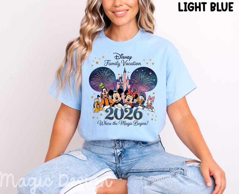 May include: A light blue t-shirt featuring a graphic of Disney characters including Mickey Mouse, Minnie Mouse, Donald Duck, Daisy Duck, Goofy, and Pluto in front of a castle with fireworks. The text reads Disney Family Vacation 2026 Where the Magic Begins.