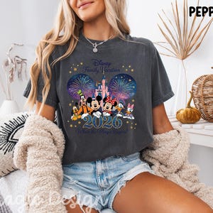 May include: A charcoal gray t-shirt featuring a graphic of Disney characters including Mickey, Minnie, Goofy, Pluto, Donald, and Daisy in front of a castle and fireworks. The text reads Disney Family Vacation 2026 Where the Magic Begins.