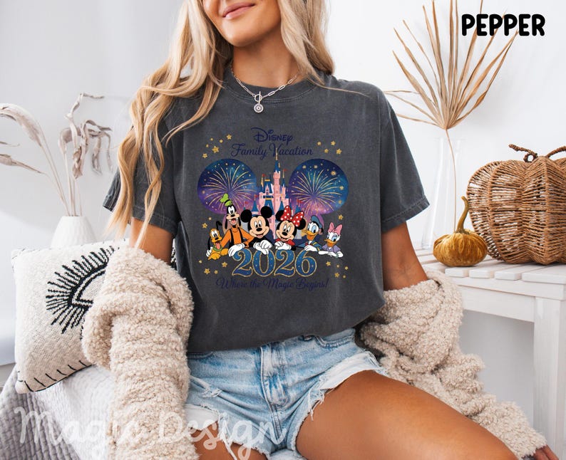 May include: A charcoal gray t-shirt featuring a graphic of Disney characters including Mickey, Minnie, Goofy, Pluto, Donald, and Daisy in front of a castle and fireworks. The text reads Disney Family Vacation 2026 Where the Magic Begins.