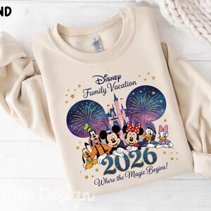 May include: A sand-colored crewneck sweatshirt featuring a graphic print of Disney characters including Mickey, Minnie, Goofy, Pluto, Donald, and Daisy. The design includes a castle, fireworks, the text Disney Family Vacation 2026 Where the Magic Begins.