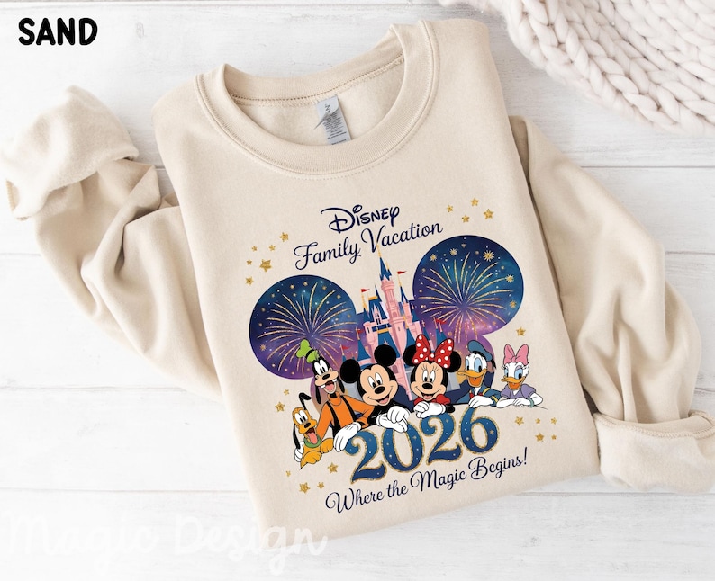 May include: A sand-colored crewneck sweatshirt featuring a graphic print of Disney characters including Mickey, Minnie, Goofy, Pluto, Donald, and Daisy. The design includes a castle, fireworks, the text Disney Family Vacation 2026 Where the Magic Begins.