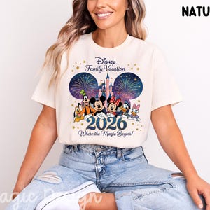 May include: A natural-colored t-shirt featuring a graphic print of Disney characters including Mickey, Minnie, Goofy, Pluto, Donald, and Daisy. The design includes a castle, fireworks, the text Disney Family Vacation, 2026, and Where the Magic Begins.
