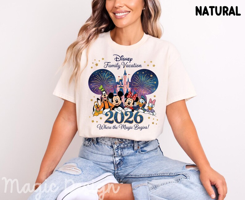 May include: A natural-colored t-shirt featuring a graphic print of Disney characters including Mickey, Minnie, Goofy, Pluto, Donald, and Daisy. The design includes a castle, fireworks, the text Disney Family Vacation, 2026, and Where the Magic Begins.