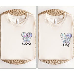 Puede incluir: Dos camisetas color crema con un dise&ntilde;o de cabeza de Mickey Mouse. Una camiseta dice "mama" y la otra "Dad". Las cabezas de Mickey Mouse est&aacute;n decoradas con un castillo y un dise&ntilde;o floral en colores pastel.