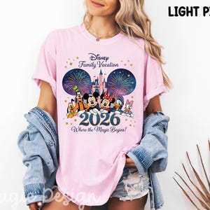 May include: A light pink t-shirt featuring a graphic print of Disney characters including Mickey Mouse, Minnie Mouse, Goofy, Donald Duck, Daisy Duck, and Pluto. The design includes a castle, fireworks, the text Disney Family Vacation 2026 Where the Magic Begins, and gold stars.
