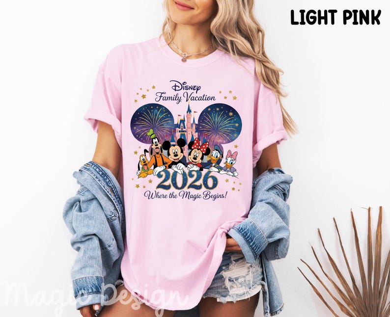 May include: A light pink t-shirt featuring a graphic print of Disney characters including Mickey Mouse, Minnie Mouse, Goofy, Donald Duck, Daisy Duck, and Pluto. The design includes a castle, fireworks, the text Disney Family Vacation 2026 Where the Magic Begins, and gold stars.