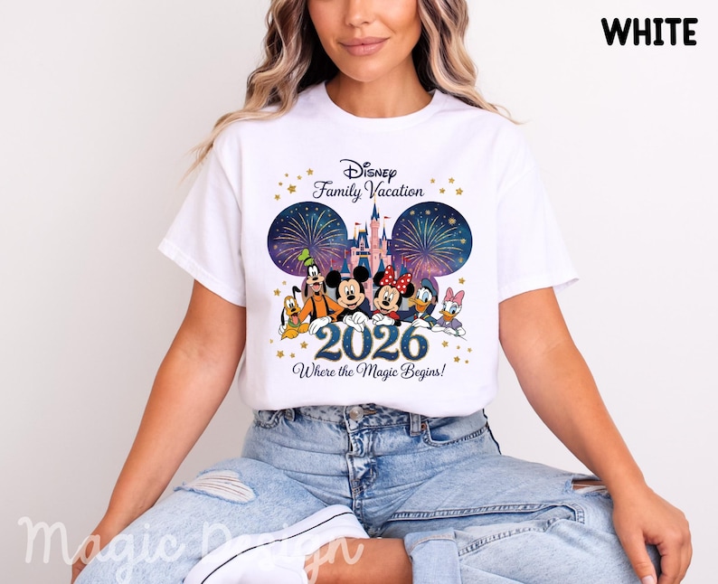 May include: A white t-shirt featuring a graphic of Disney characters including Mickey Mouse, Minnie Mouse, Goofy, Pluto, Donald Duck, and Daisy Duck in front of a castle and fireworks. The text reads Disney Family Vacation 2026 Where the Magic Begins.