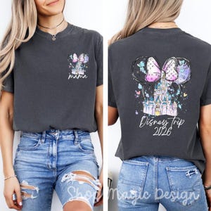 Puede incluir: Camiseta gris oscuro con un dise&ntilde;o tem&aacute;tico de Disney. La parte delantera presenta un peque&ntilde;o gr&aacute;fico con un castillo, un lazo y la palabra "mama". La parte trasera muestra orejas de Mickey Mouse, un castillo, un lazo y el texto "Disney Trip 2026."