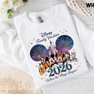 May include: A white crewneck sweatshirt featuring a graphic of Disney characters including Mickey, Minnie, Goofy, Pluto, Donald, and Daisy. The design includes a castle, fireworks, gold stars, and the text Disney Family Vacation 2026 Where the Magic Begins.