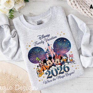 May include: An ash gray crewneck sweatshirt featuring a graphic of Disney characters including Mickey, Minnie, Donald, Daisy, Goofy, and Pluto in front of a castle with fireworks. The text reads Disney Family Vacation 2026 Where the Magic Begins.