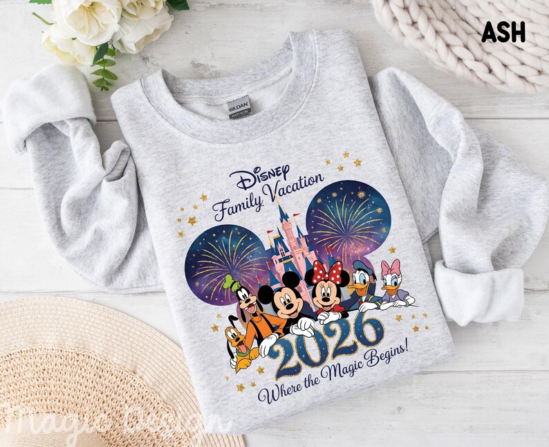 May include: An ash gray crewneck sweatshirt featuring a graphic of Disney characters including Mickey, Minnie, Donald, Daisy, Goofy, and Pluto in front of a castle with fireworks. The text reads Disney Family Vacation 2026 Where the Magic Begins.