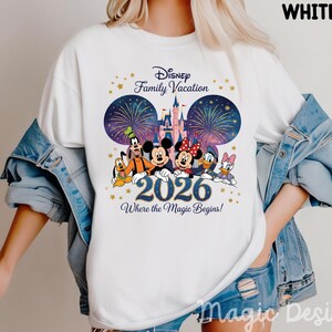May include: A white t-shirt featuring a graphic of Disney characters including Mickey, Minnie, Goofy, Pluto, Donald, and Daisy Duck in front of a castle and fireworks. The text reads Disney Family Vacation 2026 Where the Magic Begins.