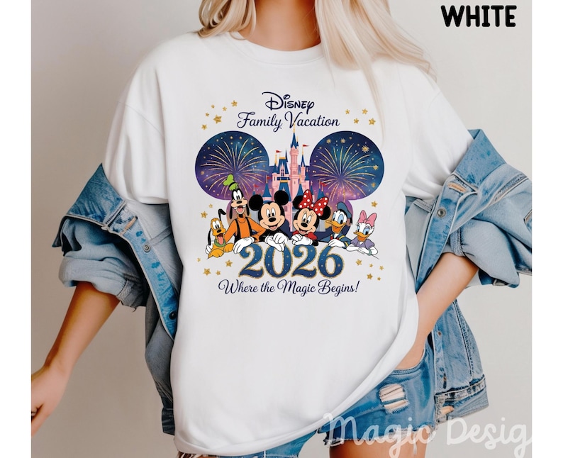 May include: A white t-shirt featuring a graphic of Disney characters including Mickey, Minnie, Goofy, Pluto, Donald, and Daisy Duck in front of a castle and fireworks. The text reads Disney Family Vacation 2026 Where the Magic Begins.