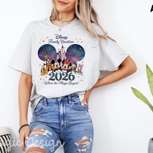May include: An ash gray short-sleeve t-shirt featuring a graphic of Disney characters including Mickey, Minnie, Goofy, Donald, and Daisy in front of a castle and fireworks. The text reads Disney Family Vacation 2026 Where the Magic Begins.