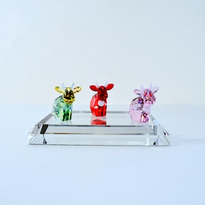 May include: Three crystal cow figurines on a clear glass base. The cows are green, red, and pink.