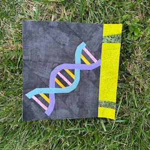 May include: A square fabric block featuring a DNA double helix design in shades of blue, purple, yellow, and pink against a dark gray background. A vertical yellow stripe with green accents is on the right side. The block is set on green grass.