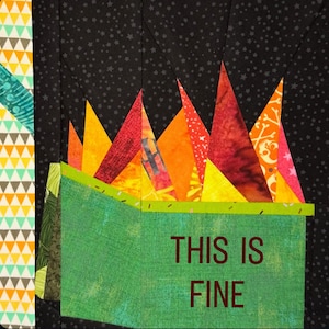 May include: A patchwork quilt block with a green box displaying the text "THIS IS FINE" in brown. Above the box are triangular shapes in red, orange, and yellow. The background is black with small white star shapes.
