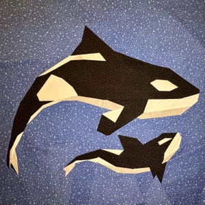 May include: A fabric art piece featuring two stylized orca whales. The whales are black and white with a geometric design, set against a blue background with white speckles. The larger whale is above the smaller one.