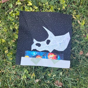 Peut inclure: Un bloc de tissu carré avec un crâne de triceratops blanc sur un fond noir parsemé d'étoiles. En dessous, des bandes de tissu avec un imprimé galaxie et le texte "He". Le bloc est posé sur de l'herbe verte.