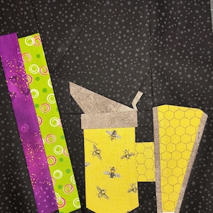 May include: A quilt block featuring a yellow mug with bee illustrations, a honeycomb design, and a gray lid. The background is black with small white stars. Vertical strips of purple and green fabric are on the left.