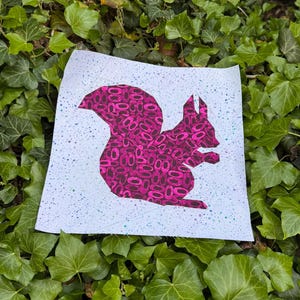 May include: A square, light blue print featuring a pink squirrel silhouette filled with a pattern of pink ovals. The background has a speckled design with blue and green accents. The print is set against a backdrop of green ivy.