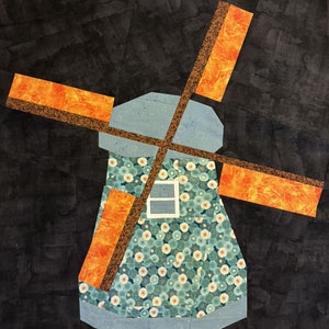 May include: A fabric windmill quilt block. The windmill features orange and brown blades, a light blue body, and a floral patterned skirt. The background is a dark gray, textured fabric.