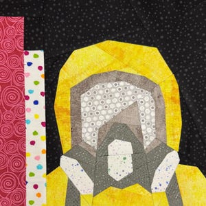 May include: A close-up of a quilt block featuring a figure with a yellow hood and gray mask. The figure is set against a black background with white stars. To the left are vertical strips of fabric with pink swirls, white polka dots, and colorful dots.