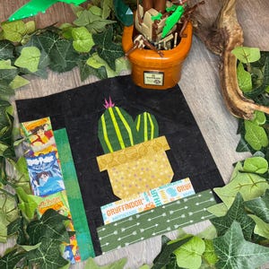 May include: A fabric block with a cactus design in a yellow pot on a black background. The design includes green and yellow accents, with strips of patterned fabric and the word "Gryffindor".