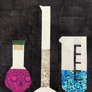 May include: A fabric art piece featuring three stylized laboratory flasks against a dark background. The flasks include a pink skull-shaped bottle, a tall white graduated cylinder, and a beaker with blue liquid. The piece is a patchwork design.