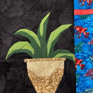 May include: A fabric art piece featuring a green plant in a tan and brown patterned pot against a dark background. The right side of the artwork shows a blue background with red, green, and purple frog illustrations.