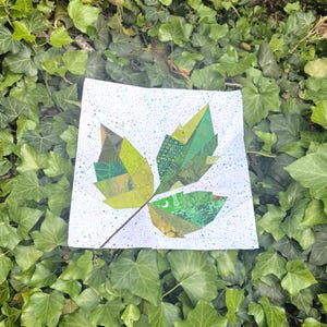 Op de afbeelding: Een vierkante, witte stof met een bladvormgeving gemaakt van verschillende groen gekleurde stoffen. Het blad heeft een bruine steel en is geplaatst tegen een achtergrond van groene klimop.