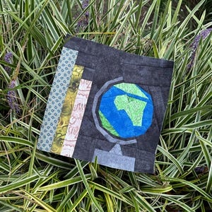 May include: A square fabric artwork depicting a globe. The globe is rendered in blue and green, set against a dark grey backdrop. The piece incorporates patterned fabric strips in blue, yellow, and pink.