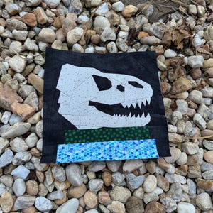 May include: A square, handmade quilt block featuring a white dinosaur skull on a black background. Below the skull are green and blue patterned fabric strips. The block is set against a background of small, multicolored stones.