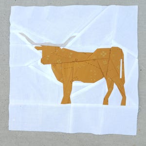 Puede incluir: Una pieza de arte textil que representa un toro longhorn en tonos dorados y blancos. El toro está hecho de tela estampada y colocado sobre un fondo blanco. El diseño geométrico es probablemente un bloque de edredón o arte textil similar.