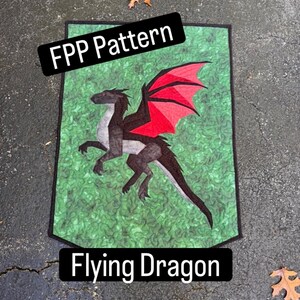 May include: A rectangular fabric wall hanging featuring a pieced quilt design of a black dragon with red wings against a green marbled background. The text FPP Pattern and Flying Dragon is overlaid on the image in black rectangular banners.