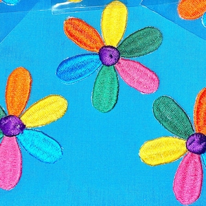 May include: Four colourful embroidered flower patches with six petals each. The petals are in shades of orange, yellow, green, blue, and pink. The centre of each flower is a purple circle.